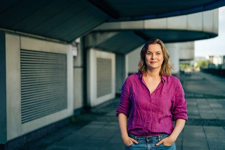 Katja Berlin mit hochgekrempelten Ärmeln und Jeans steht mit Händen in den Taschen auf einem Bürgersteig vor einem modernen Gebäude