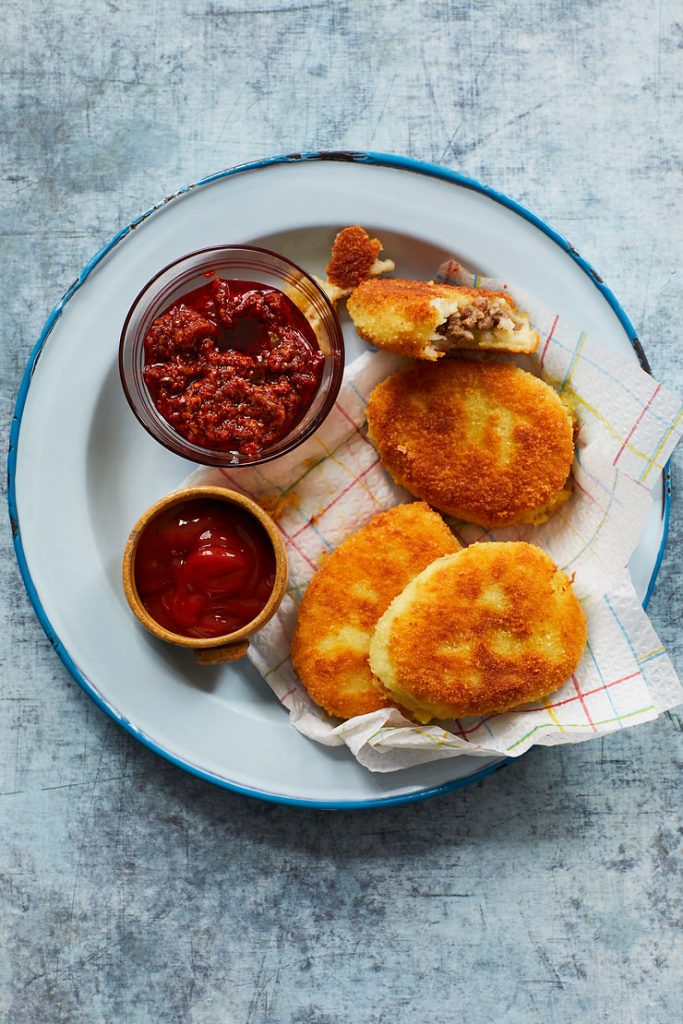 Vier panierte, goldbraun gebratene Kroketten auf einem weißen Teller mit blauem Rand, dazu zwei kleine Schälchen mit roter Sauce und Ketchup