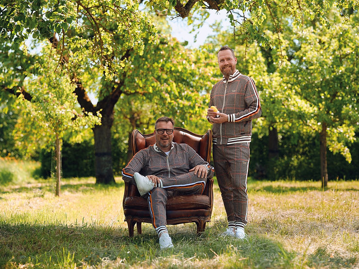 Matthias und Trent in sportlichen Freizeitanzügen, einer sitzt in einem Ledersessel, der andere steht daneben in einem Garten mit Bäumen im Hintergrund