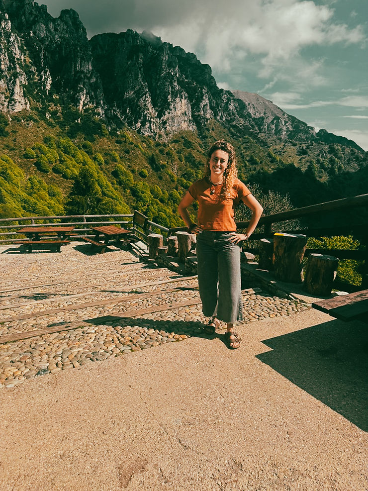 Frau mit Zopf, orangefarbenem T-Shirt und grauer Hose steht auf gepflastertem Platz vor bewaldetem Bergmassiv