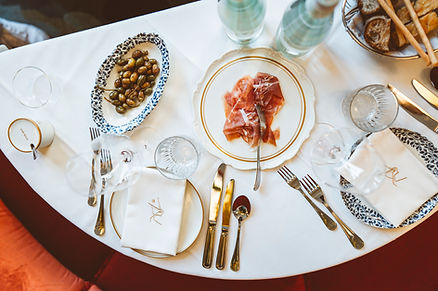 Gedeckter Tisch mit Tellern, Besteck, Gläsern, Servietten und einer Platte mit Schinken und einer Schale mit Oliven