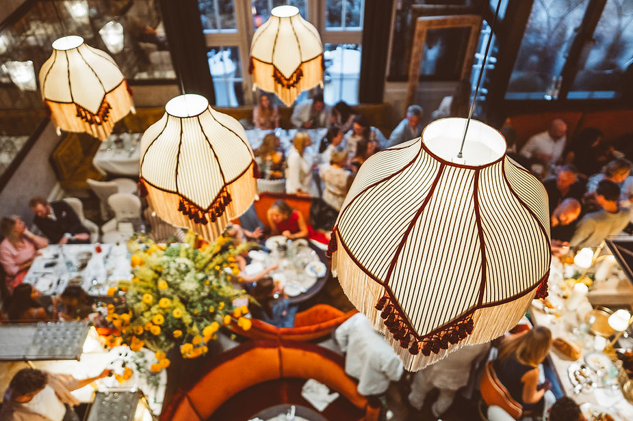 Von oben sichtbare, große, dekorative Lampenschirme über einem belebten Restaurant mit mehreren Gästen und einem runden Sofa mit Blumenarrangement in der Mitte