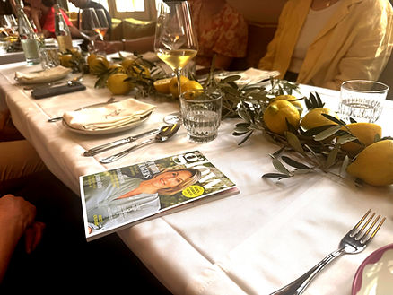 Tisch mit weißer Tischdecke, darauf liegen Gläser, Besteck, Zitronen mit Zweigen und ein Magazin mit einem Porträt auf dem Cover