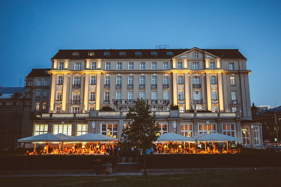 Beleuchtetes mehrstöckiges historisches Gebäude mit großen Fenstern und Außenbereich mit Sonnenschirmen und Menschen am Abend
