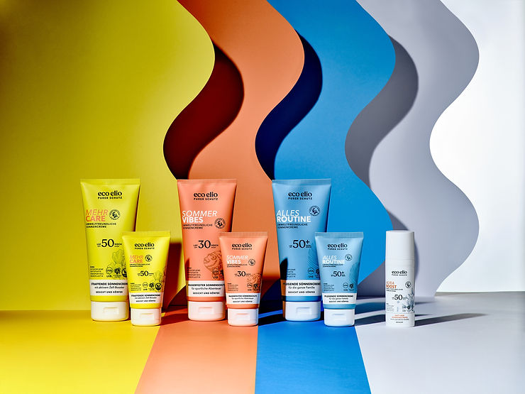 Mehrere Sonnencremeprodukte der Marke eco ello in gelben, orangen, blauen und weißen Verpackungen vor farblich passenden wellenförmigen Hintergründen