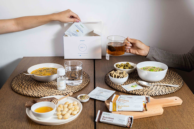 Zwei Personen an einem Tisch mit verschiedenen Snacks, Getränken und einer geöffneten weißen Box mit der Aufschrift 'DAY 1'