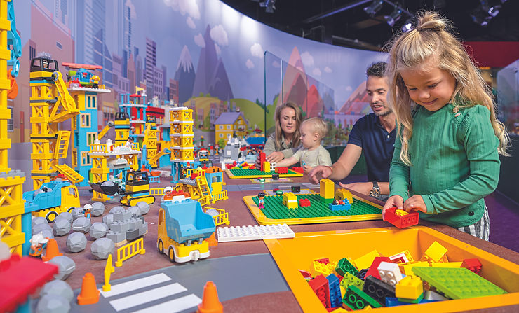 Kinder spielen an einem Tisch mit bunten Bausteinen vor einer großen Wand mit einer Stadt- und Berglandschaft als Hintergrund
