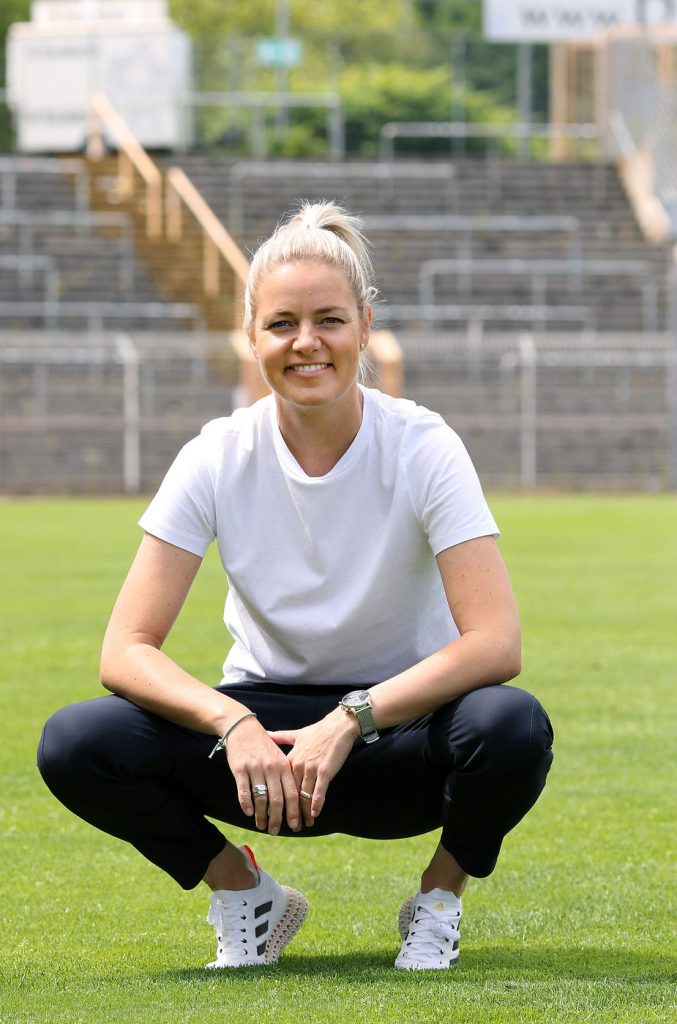Christina Graf mit weißem T-Shirt, schwarzer Hose und weißen Sportschuhen hockt auf einem Sportplatz