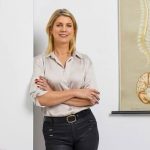 Frau mit verschränkten Armen lehnt an einer weißen Wand neben einem medizinischen Poster mit Wirbelsäule und Gehirn