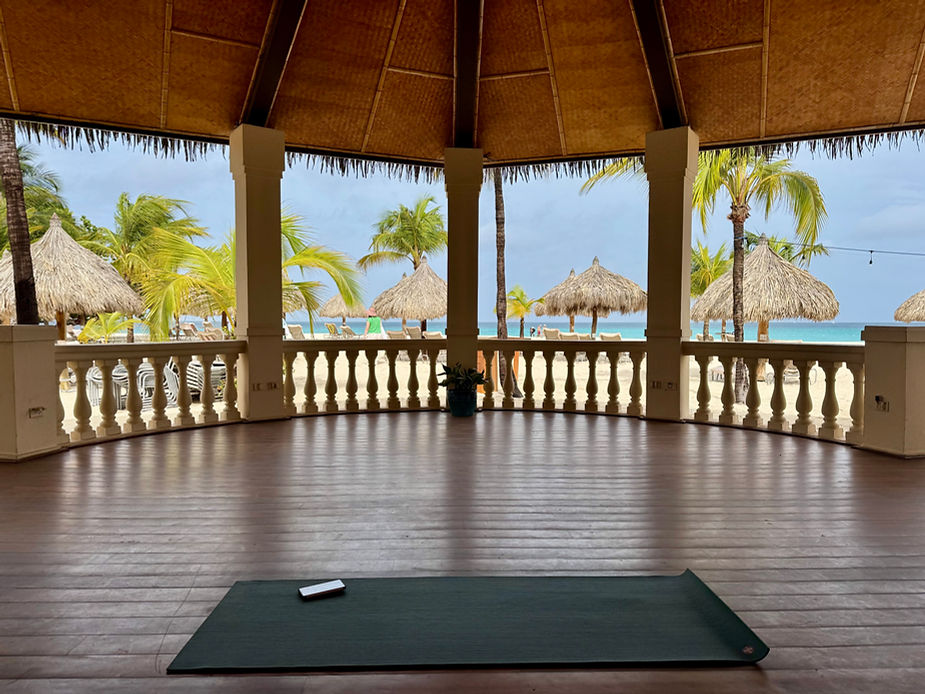 Überdachte Terrasse mit Säulen und balustradenförmigem Geländer, Blick auf Palmen und Strohdachhütten am Strand, auf dem Boden liegt eine Yogamatte mit einem Smartphone