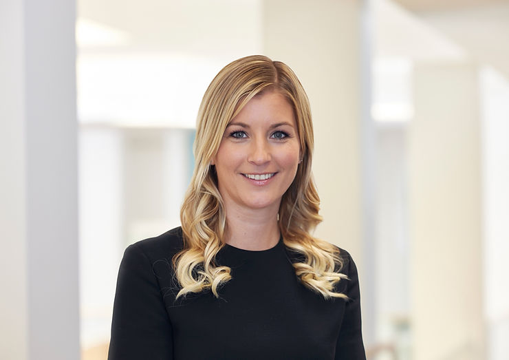 Frau mit langen, gewellten blonden Haaren trägt ein schwarzes Oberteil und steht vor einem hellen, unscharfen Hintergrund