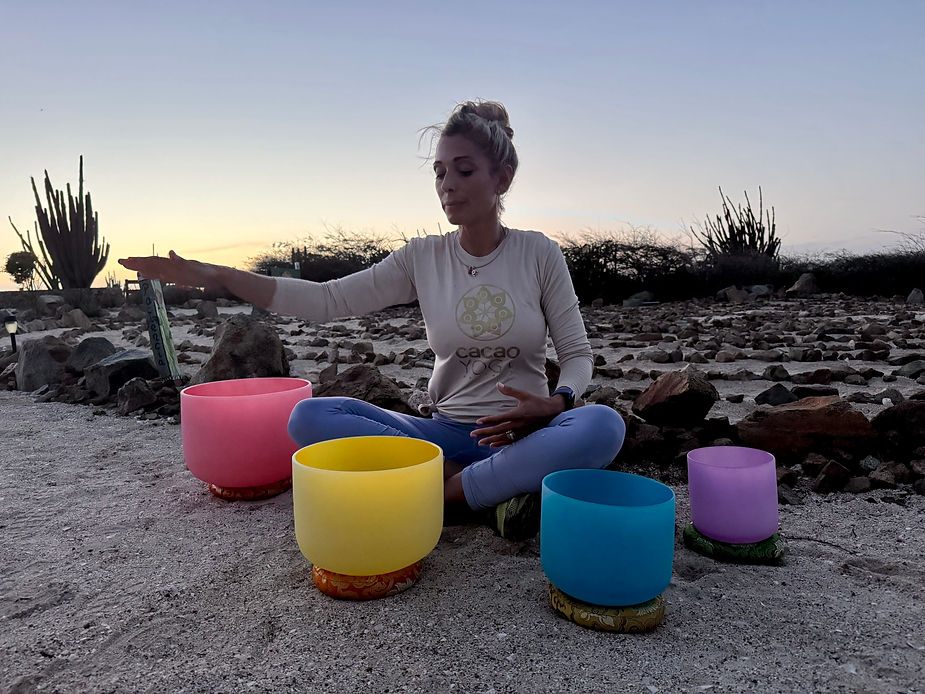 Person sitzt im Schneidersitz auf sandigem Boden vor einer Anordnung von vier farbigen Klangschalen und streckt einen Arm seitlich aus