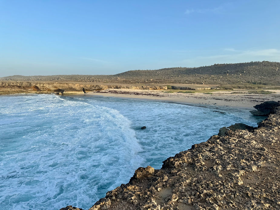 Küstenlandschaft mit felsigem Vordergrund, Brandung und flachem Strand vor hügeliger, karger Vegetation unter klarem Himmel