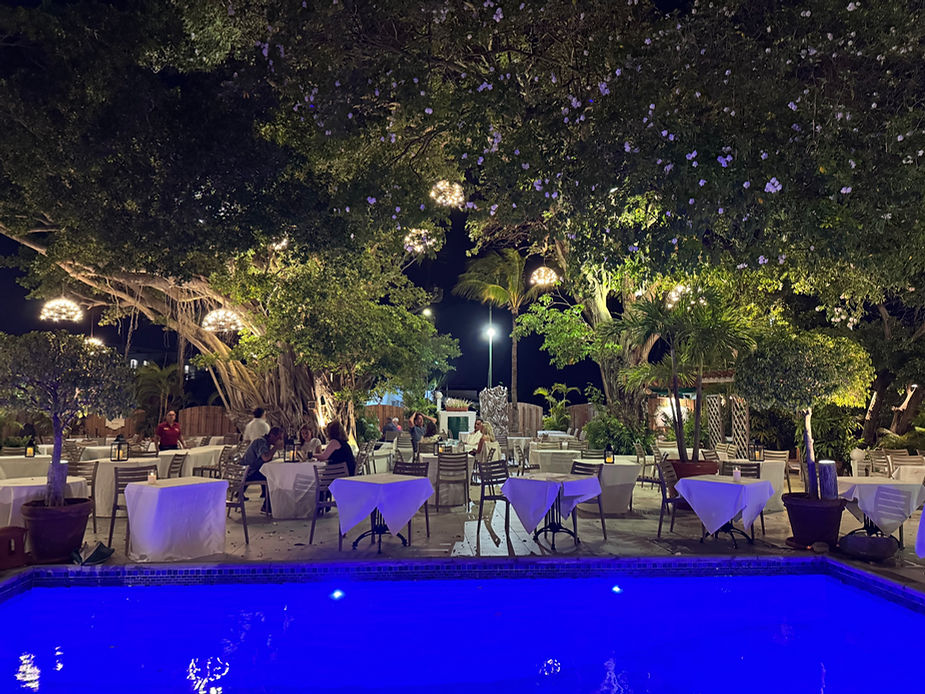 Außenrestaurant mit gedeckten Tischen unter großen Bäumen neben einem beleuchteten Pool bei Nacht