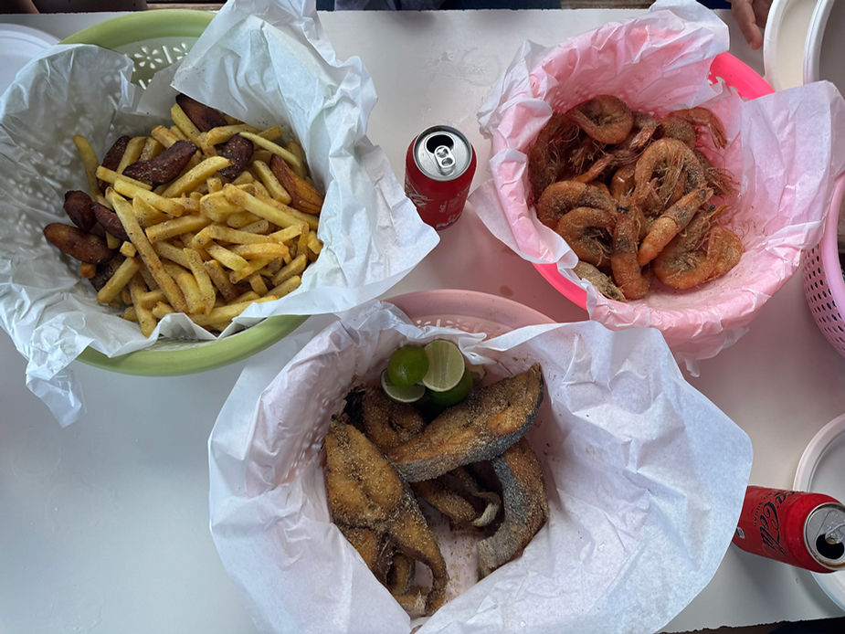 Drei Schüsseln mit frittierten Pommes und Würstchen, Garnelen und gebratenem Fisch mit Limettenscheiben auf einem weißen Tisch mit zwei Dosen Cola