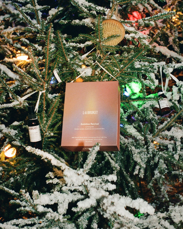 Weihnachtsbaum mit Kunstschnee und mehreren hängenden Kosmetikfläschchen sowie einer braunen Verpackung mit der Aufschrift 'L:A BRUKET Solstice Revival'