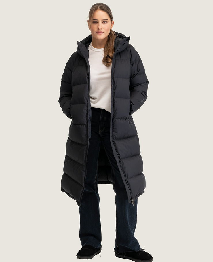 Frau trägt lange, schwarze Steppjacke mit Kapuze, weiße Bluse, dunkle Jeans und schwarze Schuhe