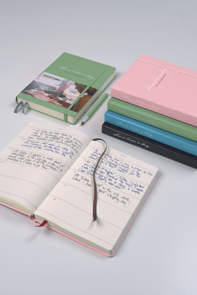 Aufgeschlagenes Notizbuch mit handschriftlichen Einträgen und Lesezeichen neben mehreren geschlossenen Notizbüchern in verschiedenen Farben und zwei Stiften