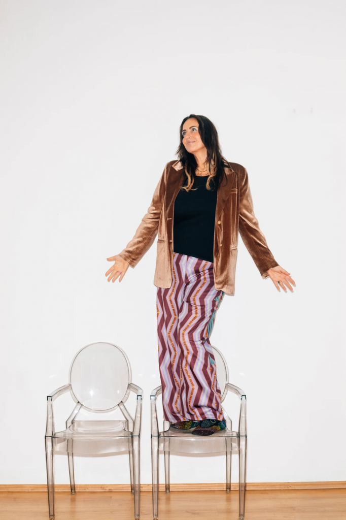 Sandra Günther steht mit ausgestreckten Armen auf einem transparenten Stuhl vor weißer Wand und trägt eine braune Jacke und eine gemusterte lange Hose