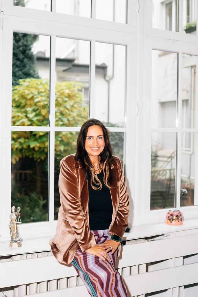 Sandra Günther mit langen dunklen Haaren trägt schwarzen Pullover, rosafarbenen Samtblazer und gestreifte Hose und sitzt auf einer Fensterbank vor großen Fenstern mit Blick auf einen Garten.