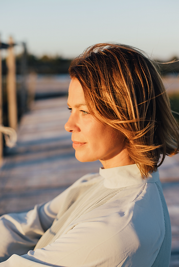 Jessica Schwarz mit schulterlangem, braunem Haar sitzt seitlich auf einem Holzsteg im Freien