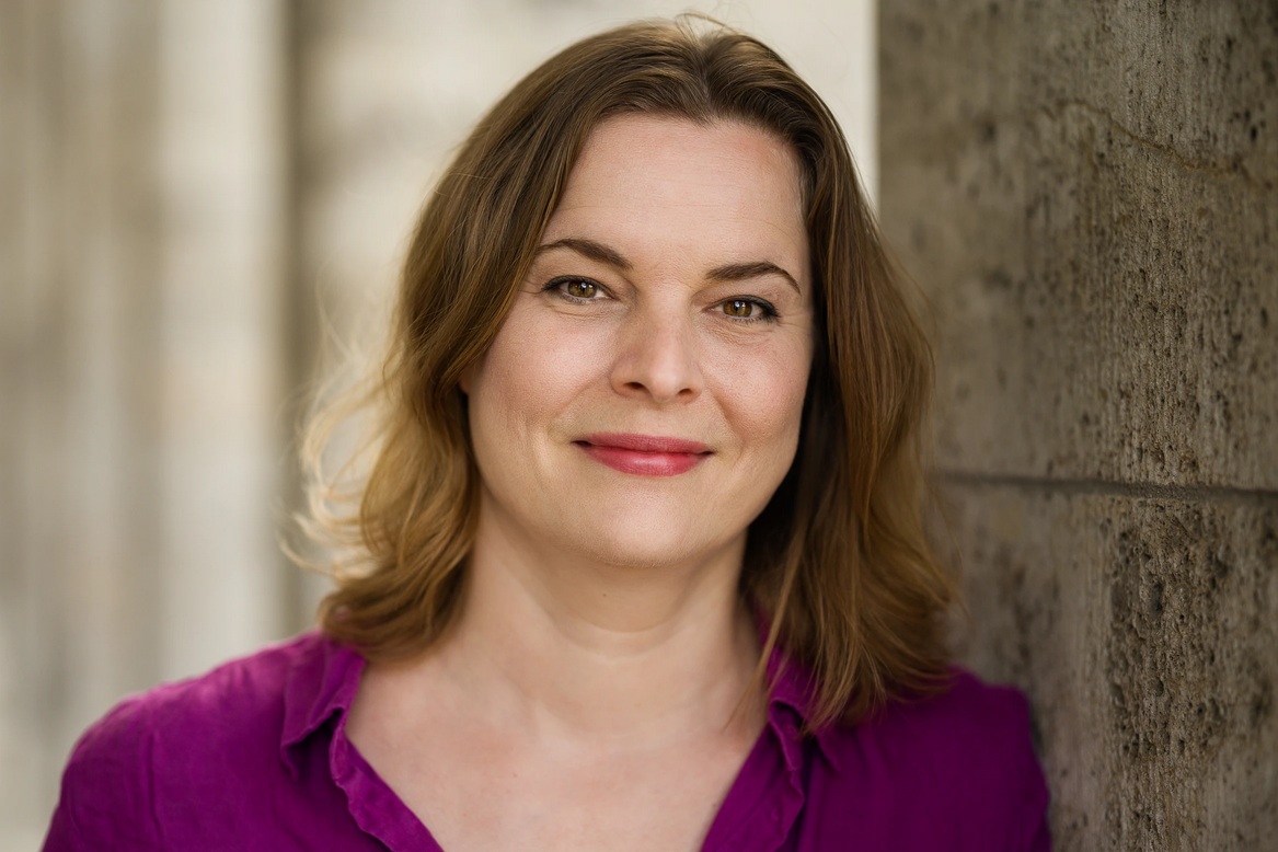 Katja Berlin mit schulterlangen, gewellten Haaren trägt ein purpurfarbenes Hemd und lehnt an einer hellen Wand