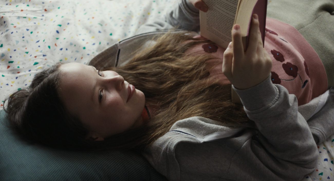 Mädchen (Marielle/Laeni Geiseler) liegt auf dem Rücken auf einem Bett mit weißer Bettwäsche und buntem Muster und liest ein Buch