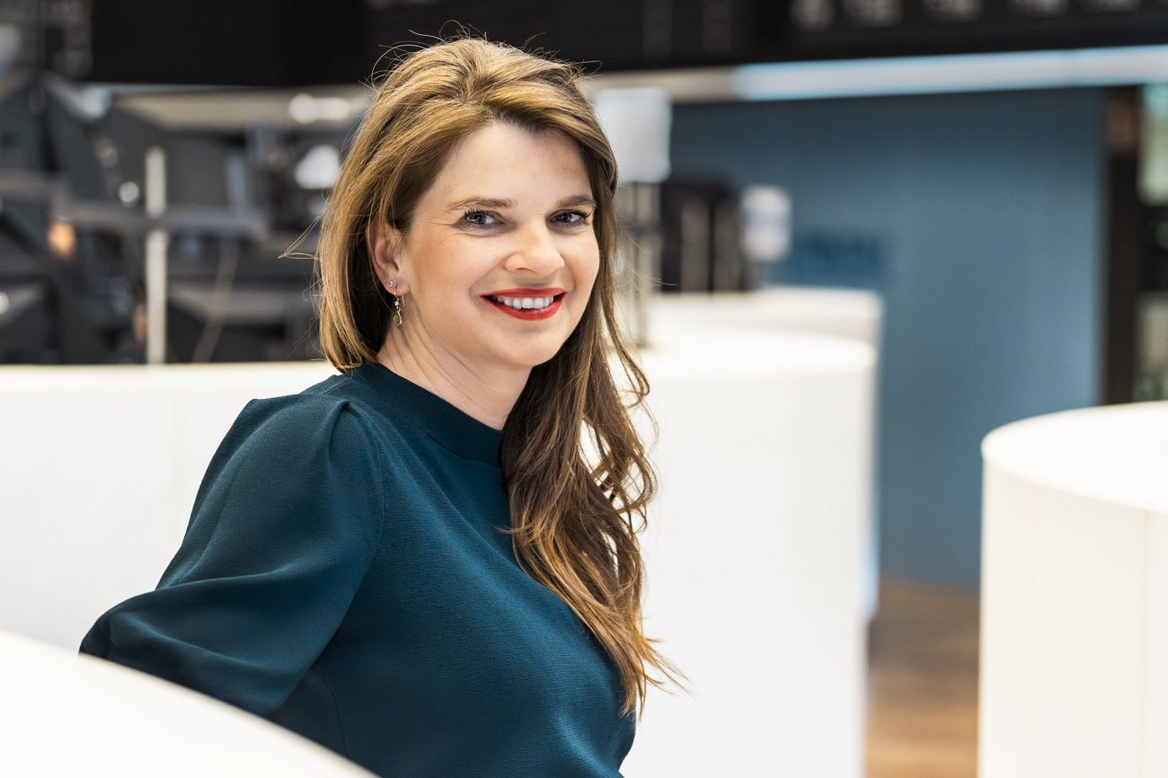 Frau mit langen, gewellten Haaren sitzt in einem modernen Innenraum auf einem weißen Sofa und trägt einen dunkelgrünen Pullover