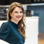 Frau mit langen, gewellten Haaren sitzt in einem modernen Innenraum auf einem weißen Sofa und trägt einen dunkelgrünen Pullover