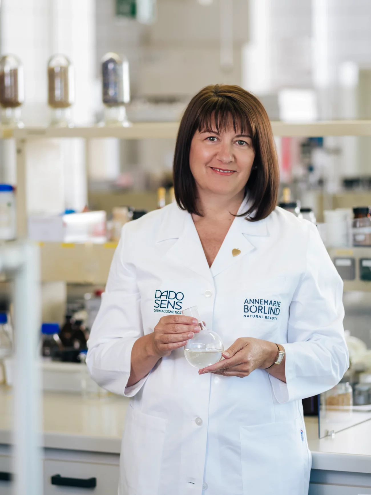 Frau in weißem Laborkittel mit den Aufschriften 'DADO SENS' und 'ANNEMARIE BÖRLIND NATURAL BEAUTY' hält ein rundes Glasgefäß in einem Labor.