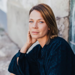 Jessica Schwarz mit schulterlangem Haar lehnt mit dem Ellenbogen an einer Wand und trägt eine dunkelblaue Bluse