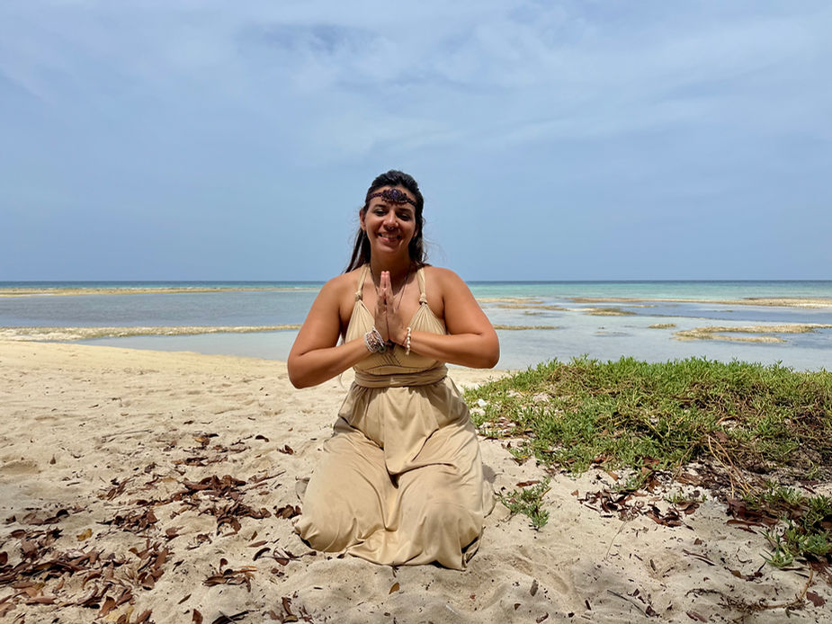 Person kniet im Sand am Strand mit gefalteten Händen vor dem Oberkörper und trägt ein ärmelloses Oberteil und eine weite Hose