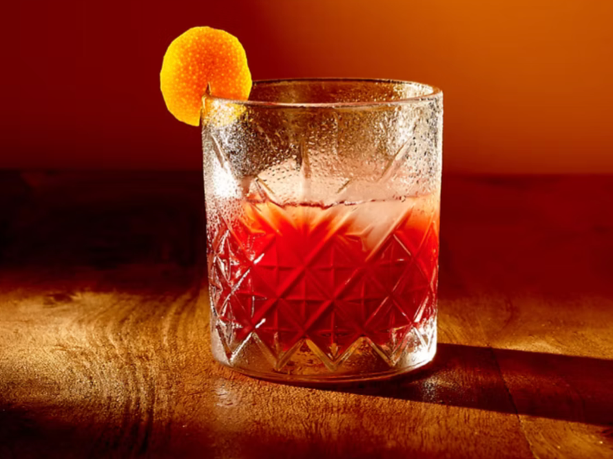 Kristallglas mit rotem Getränk und Orangenscheibe am Glasrand auf Holztisch vor orangefarbenem Hintergrund