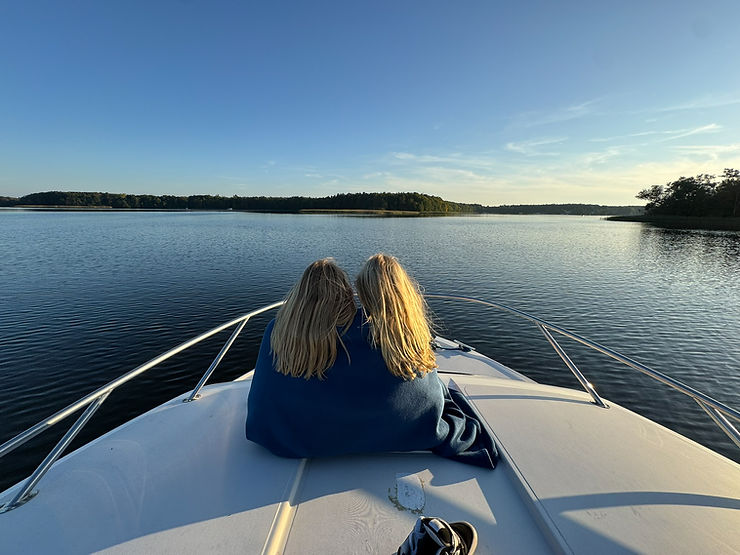 Zwei Mädchen mit langen blonden Haaren sitzen auf dem Bug eines Bootes und blicken auf einen ruhigen See mit bewaldetem Ufer