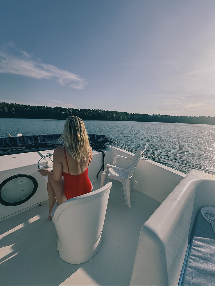 Frau mit rotem Badeanzug sitzt auf einem weißen Stuhl auf einem Boot und blickt auf einen See mit bewaldetem Ufer