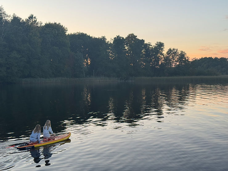 Zwei Personen sitzen auf einem Kajak auf einem ruhigen See bei Sonnenuntergang mit Bäumen am Ufer