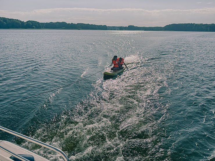 Zwei Personen mit Schwimmwesten sitzen in einem Kajak auf einem See, das Kajak wird von einem Boot gezogen