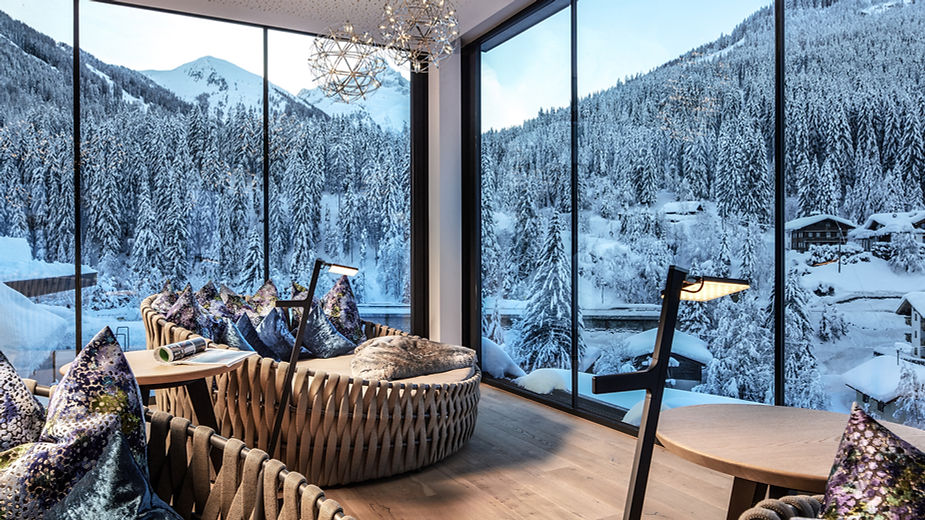 Wohnzimmer mit großen Panoramafenstern, zwei runden Sofas mit Kissen und zwei Stehlampen vor verschneiter Berglandschaft