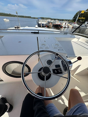 Blick von einem Boot auf das Steuerrad mit zwei Personen, die Beine sind sichtbar, im Hintergrund Wasser und weitere Boote am Steg