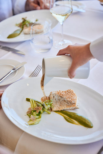 Hand gießt Sauce aus einem weißen Gefäß auf ein kunstvoll angerichtetes Fischgericht mit grünem Gemüse auf weißem Teller