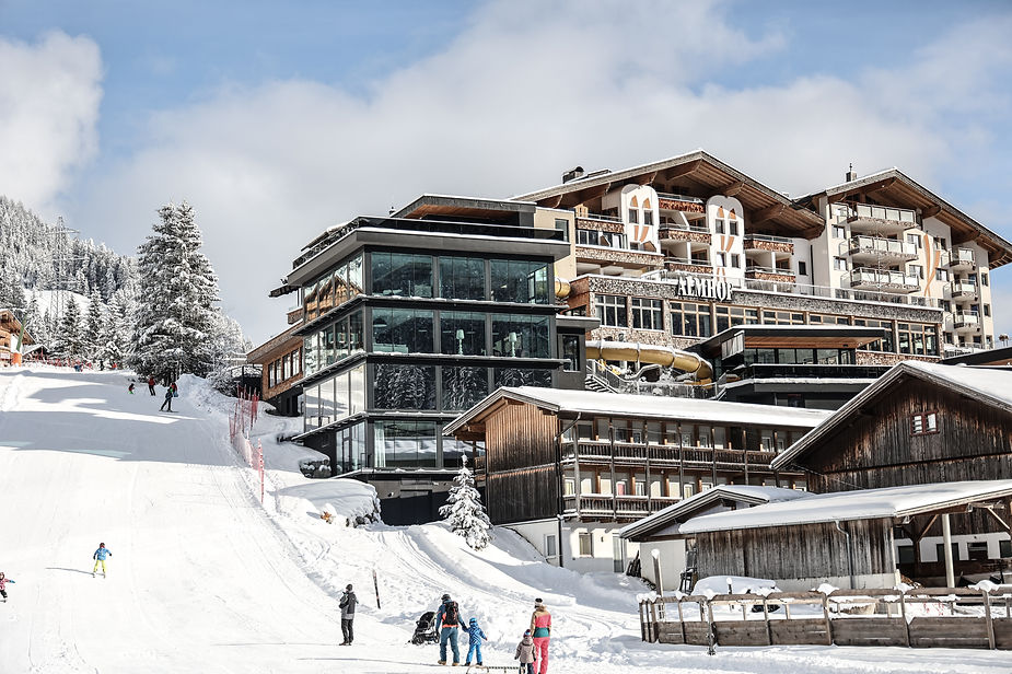 Skigebiet mit mehreren Gebäuden, darunter ein modernes Glasgebäude und traditionelle Holzhäuser, auf schneebedecktem Hang mit Skifahrern