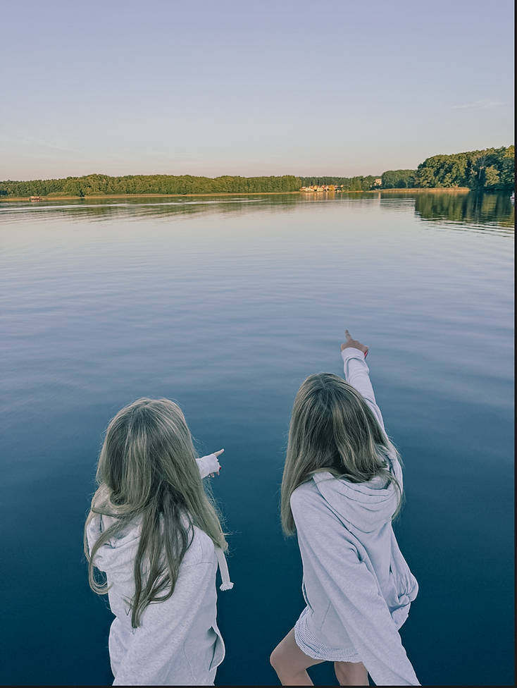 Zwei Mädchen mit hellen Kapuzenjacken zeigen auf einen See mit bewaldetem Ufer im Hintergrund