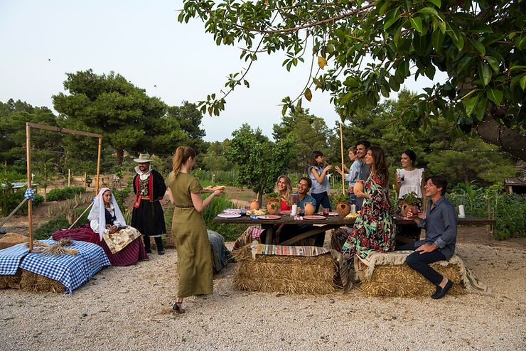 Gruppe von Menschen sitzt und steht an einem Holztisch mit Strohballen als Sitzgelegenheiten in einem Garten mit Bäumen und Kiesboden