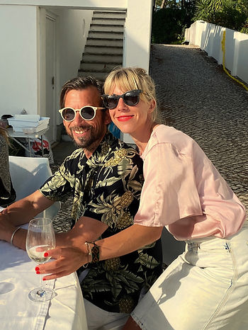 Sara Devi Graaf und Nikolaus Ronacher sitzen an einem Tisch im Freien, sie trägt ein helles Hemd und einen weißen Rock, er ein dunkles Hemd mit floralem Muster, auf dem Tisch steht ein Weinglas.