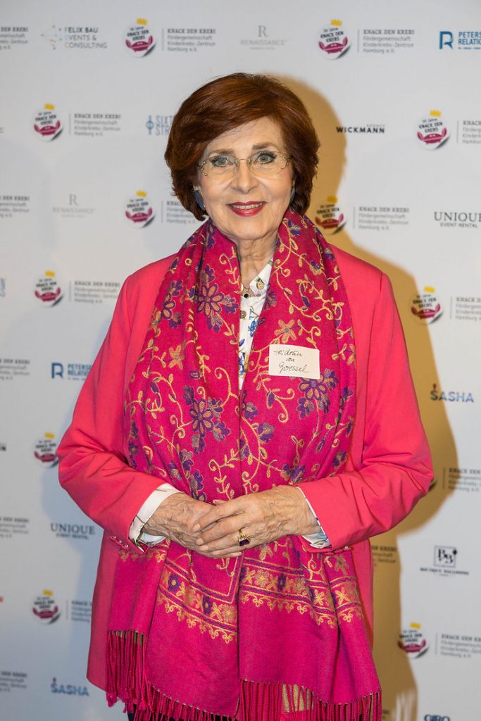 Heidrun von Goessel mit rotem Blazer und pink-rotem Schal vor einer Wand mit Logos und Texten