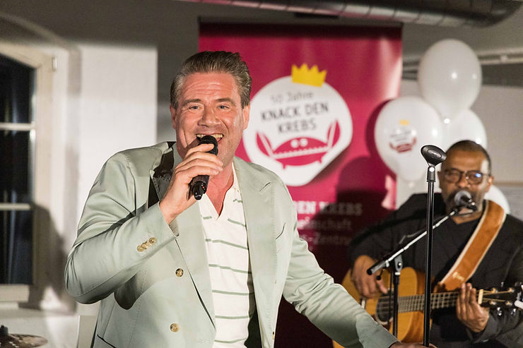 Sasha in hellgrauem Sakko hält Mikrofon vor rotem Banner mit Krone und Text, im Hintergrund Person mit Gitarre und weißen Luftballons