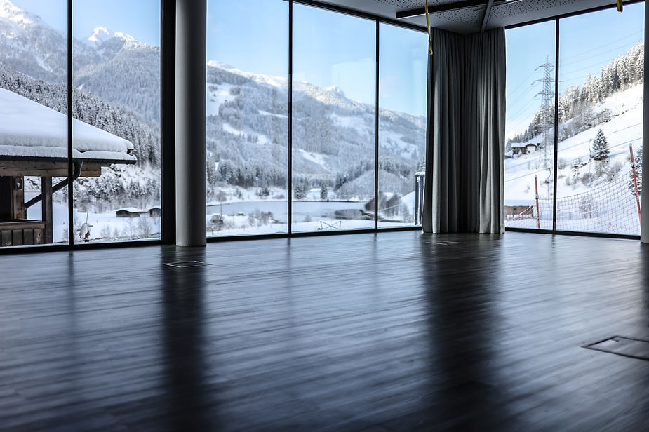 Großer leerer Raum mit dunklem Holzboden und bodentiefen Fenstern mit Blick auf schneebedeckte Berge und Häuser