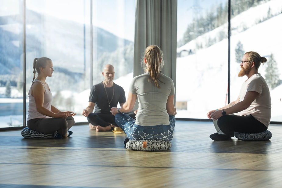 Vier Personen sitzen im Schneidersitz auf Meditationskissen in einem Raum mit großen Fenstern und Bergblick