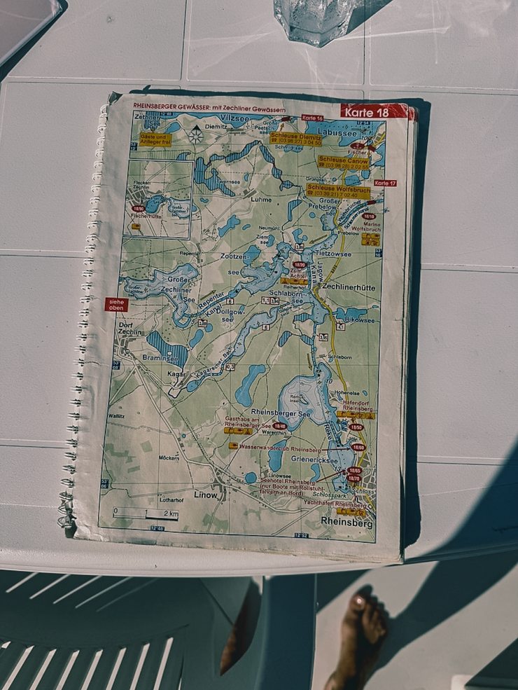 Karte mit Gewässern und Orten rund um Rheinsberg auf einem Spiralblock auf weißem Tisch mit Schatten und nackten Füßen im Bild