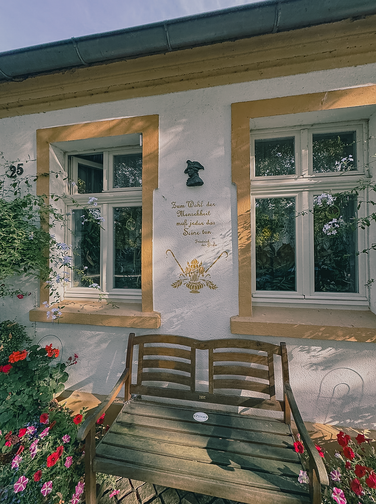 Hausfassade mit zwei Fenstern, einer Holzbank davor und einer goldfarbenen Schrift mit Zitat zwischen den Fenstern